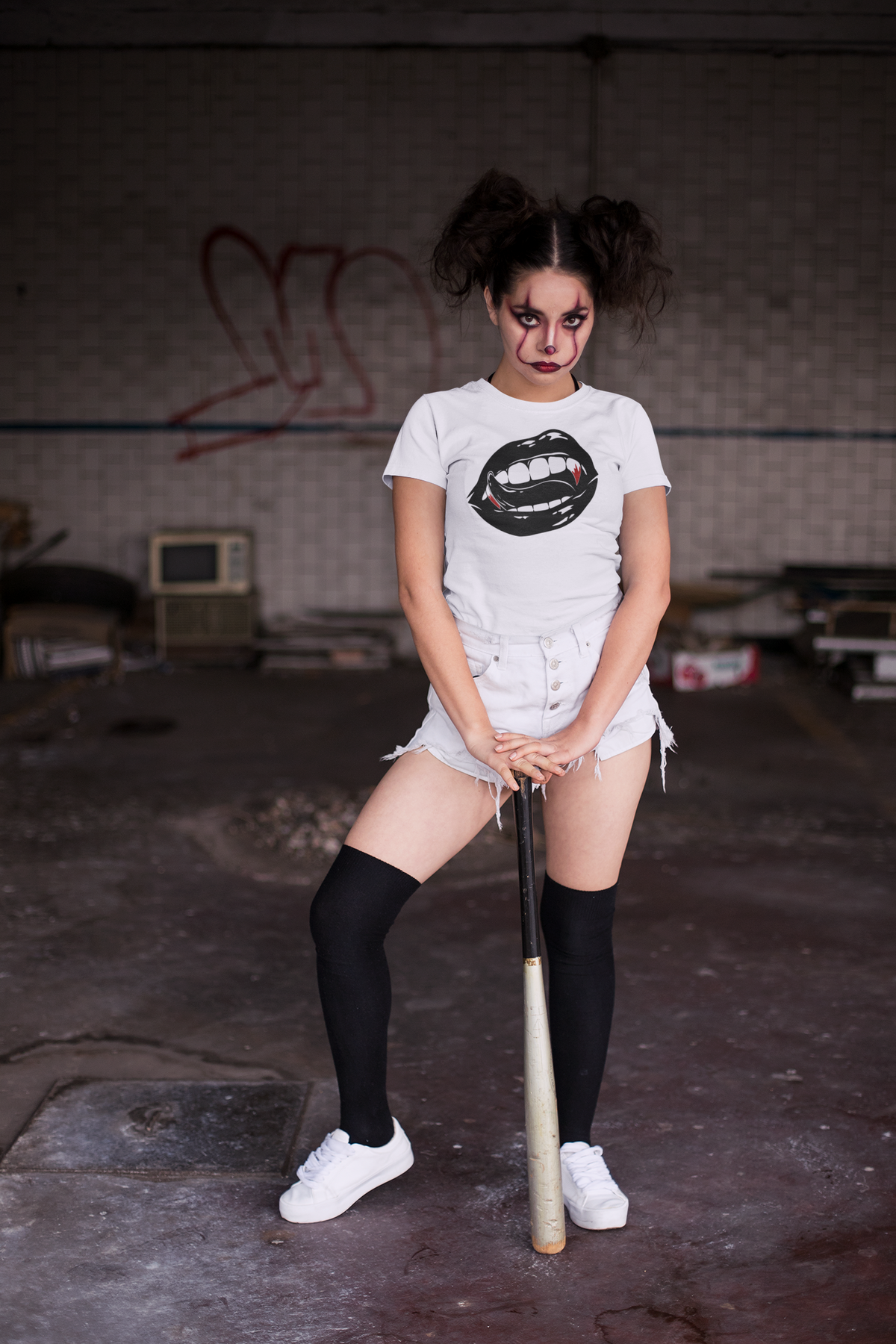 A woman with dark hair in a messy bun holds a baseball bat over her shoulder, wearing a black graphic t-shirt with a white and black lips design, white denim shorts, and black knee-high socks, set against a solid dark mauve background.
